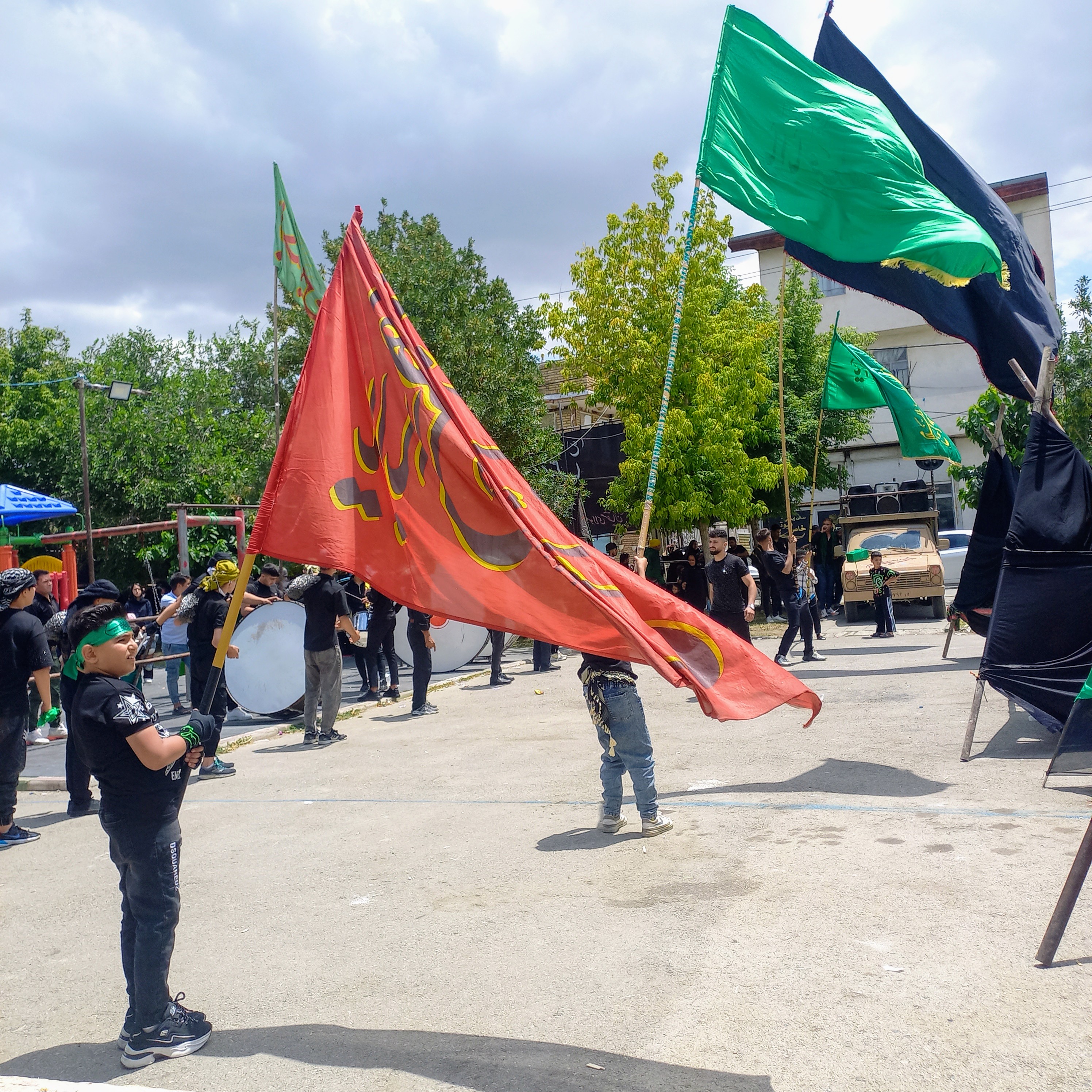 پرچم بزرگ سرخ «یا ابالفضل العباس » در دستان کوچک یک کودک سیاه‌پوش به اهتزاز درآمده و باد تاسوعا آن را همچون شعله‌ای فروزان به رقص آورده است. پشت سرش طبل‌های عزا می‌نوازند و عزاداران با شور و اشک، یاد خون خدا را زنده نگه داشته‌اند.تاسوعای حسینی | ۲۵ تیر ۱۴۰۳ | ارومیه، بوستان صفا 🖤 یا لثارات الحسین