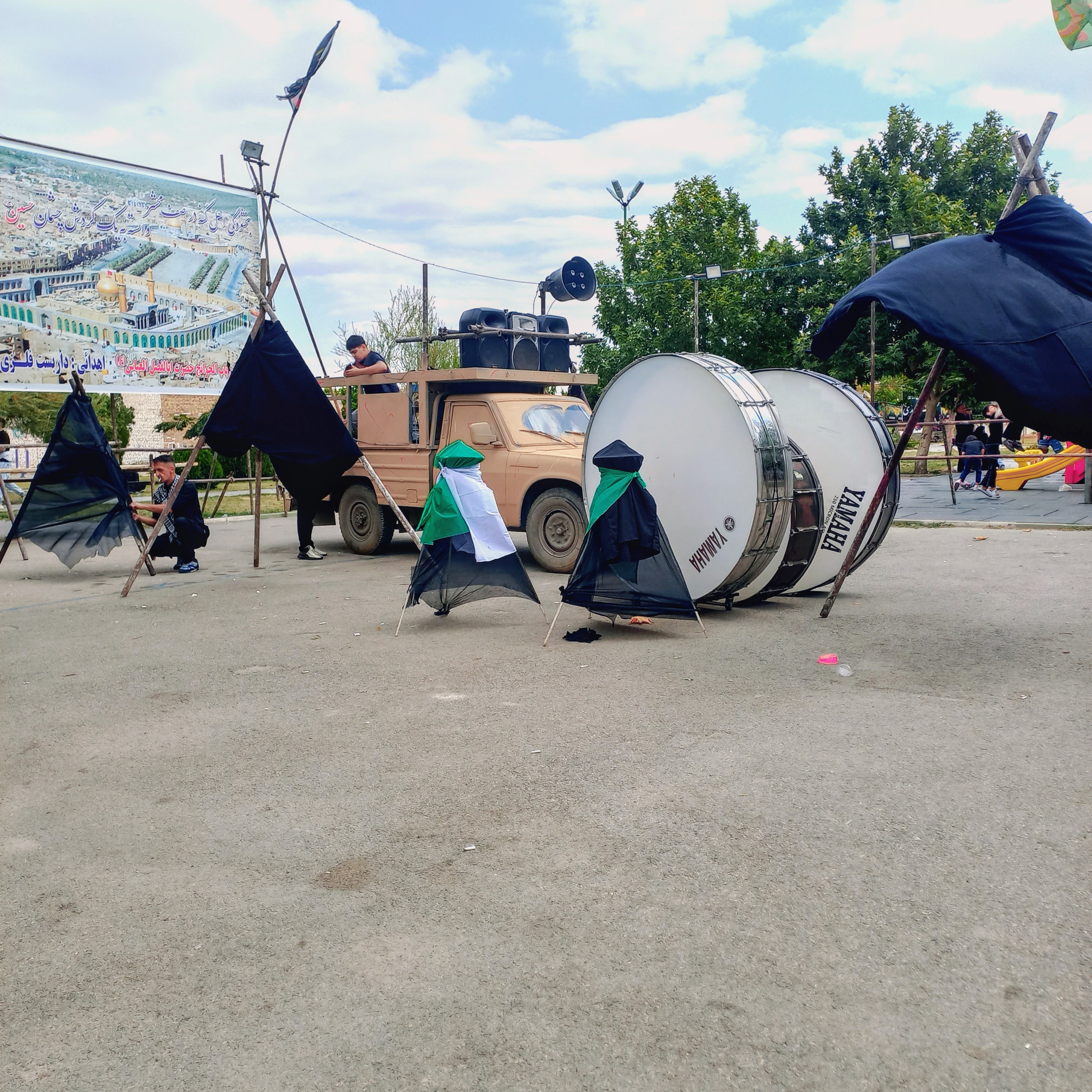 طبل بزرگ عزا در روز عاشورا بر جلوی وانت قرار گرفته و آماده نواختن نوای «حسین حسین» است. علامت‌های سیاه‌پوش با پرچم‌های سبز بر دوش کودکان و نوجوانان سنگینی می‌کند و بنر بزرگ عزاداری در پس‌زمینه نصب شده؛ این قاب، نشان‌دهنده شور و حال عزاداری حسینی در ارومیه است که با صدای طبل و اشک عزاداران، یاد کربلا را زنده می‌کند.عاشورا حسینی | ۲۶ تیر ۱۴۰۳ | ارومیه، بوستان صفا 🖤 یا حسین (ع)