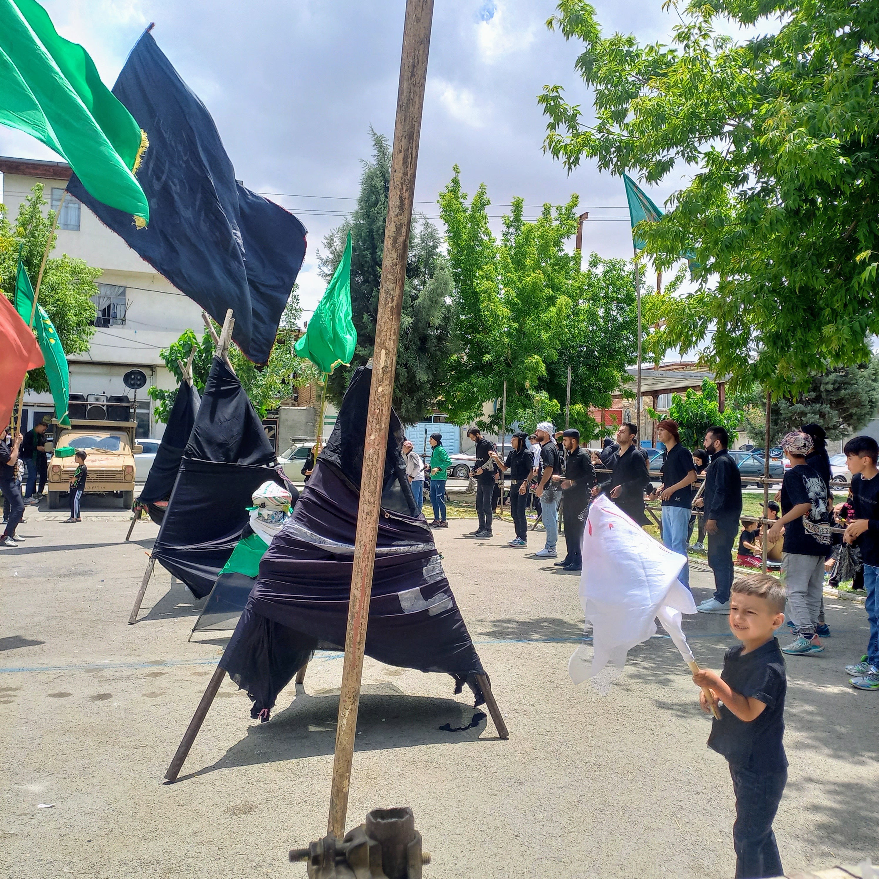 علامت‌های سنگین سیاه‌پوش عزا، نماد ماتم اباعبدالله (ع)، پیشاپیش دسته حرکت می‌کنند و پرچم‌های سبز و سیاه در نسیم تاسوعا به اهتزاز درمی‌آیند. کودکی با لبخند معصومانه و پرچمی سفید در دست، جلوه‌ای از عشق خالصانه نسل نو به حسین (ع) است.تاسوعای حسینی | ۲۵ تیر ۱۴۰۳ | ارومیه، بوستان صفا 🖤 یا اباعبدالله(ع)