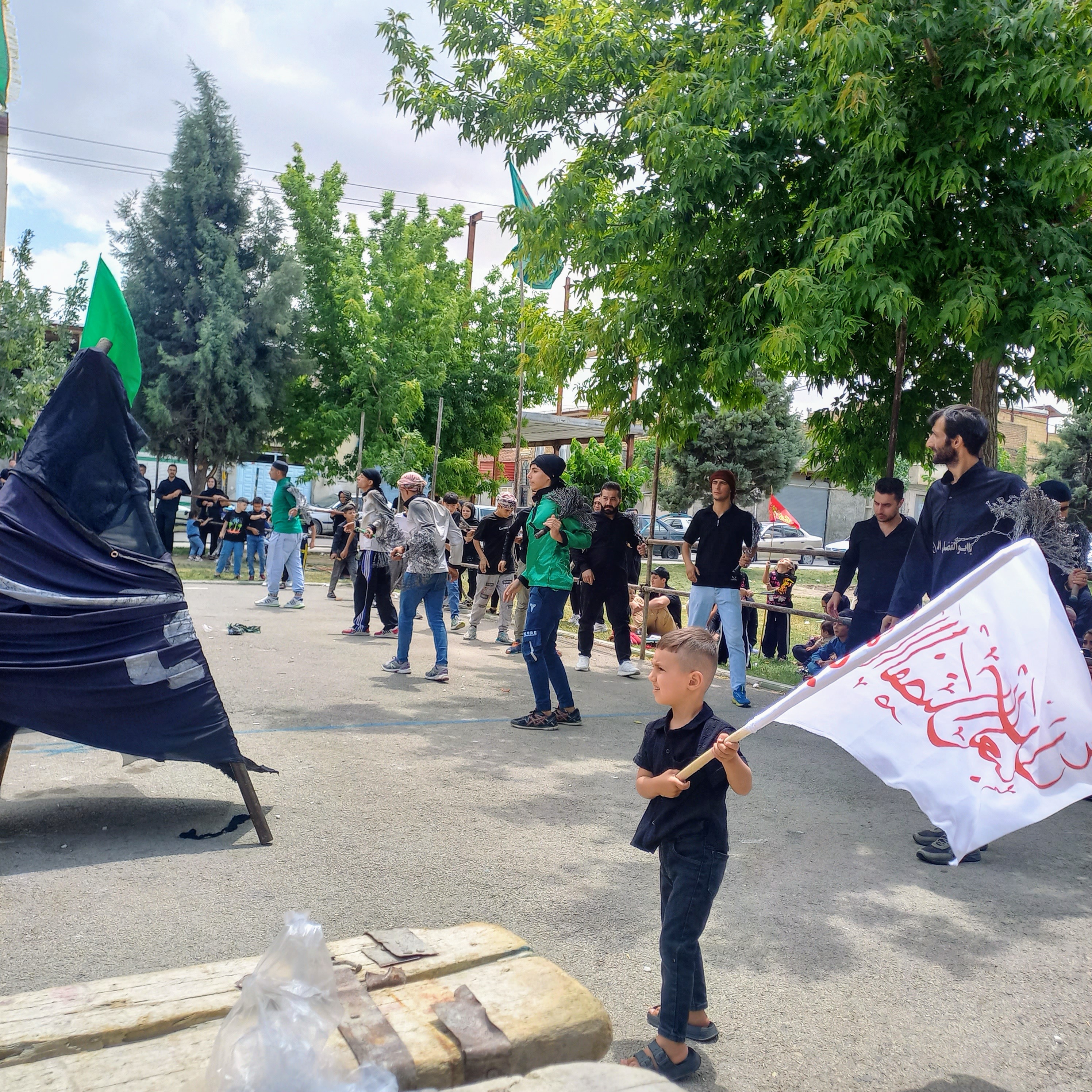 کودکی معصوم با پرچمی سفید و نوشته‌ای سرخ بر دوش، در میان عزاداران سیاه‌پوش گام برمی‌دارد: «یا ابالفضل العباس ». علامت‌های سنگین عزا و پرچم‌های سبز در پیشاپیش دسته حرکت می‌کنند و این صحنه، گواه آن است که نام حسین (ع) از کودکی بر دل‌ها حک شده و تا ابد ماندگار خواهد ماند. تاسوعای حسینی | ۲۵ تیر ۱۴۰۳ | ارومیه، بوستان صفا 🖤 یا اباعبدالله الحسین