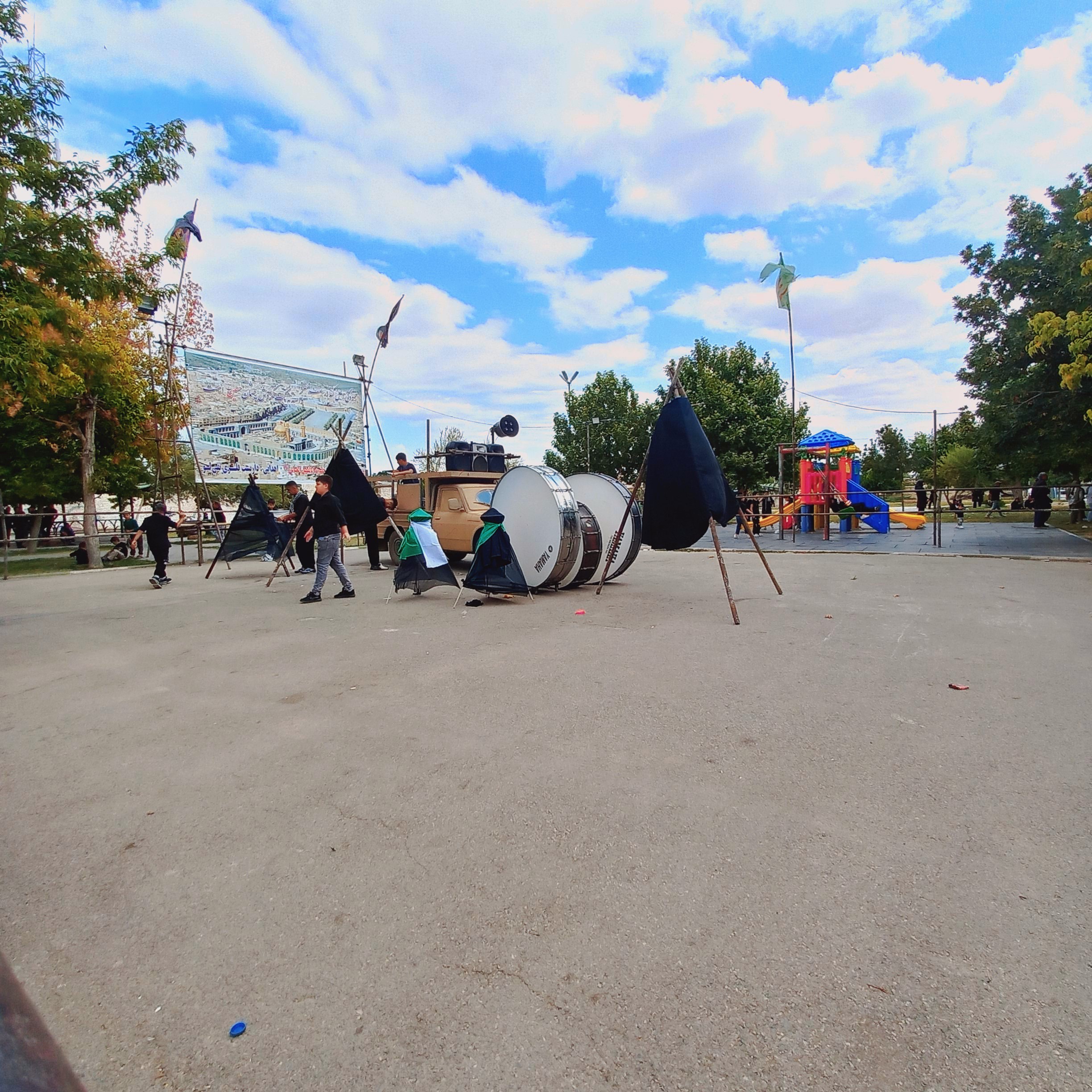 در روز عاشورا، آماده‌سازی دسته عزاداری در بوستان صفا به اوج رسیده؛ طبل‌های بزرگ عزا بر زمین قرار گرفته، علامت‌های سیاه‌پوش برپا شده و بنر بزرگ با تصویر حرم امام حسین (ع) نصب گردیده است. کودکان و نوجوانان سیاه‌پوش با شور حسینی دور هم جمع شده‌اند تا نوای ماتم کربلا را بلند کنند؛ این لحظه، آغاز فریاد «یا حسین» از دل‌های عاشق است.عاشورا حسینی | ۲۶ تیر ۱۴۰۳ | ارومیه، بوستان صفا 🖤  یا حسین(ع)