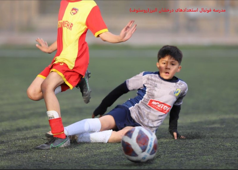 تصویر هیجان انگیز از بازی فوتبال دو نوجوان در زمین چمن یکی با لباس طوسی و آرم روستوف ایران و دیگری با لباس قرمز